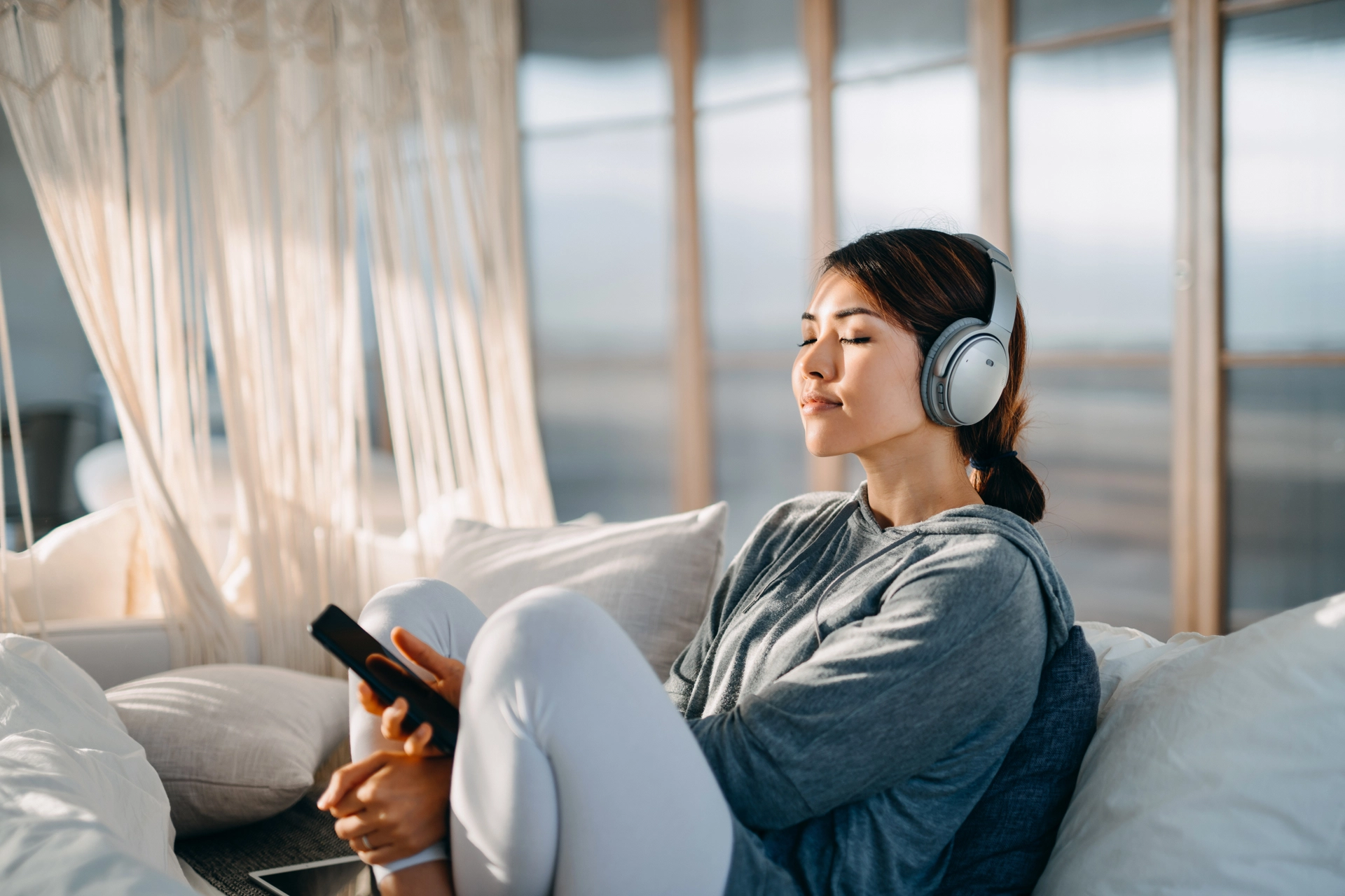 Relaxed woman with eyes closed sitting on bed enjoying music over headphones